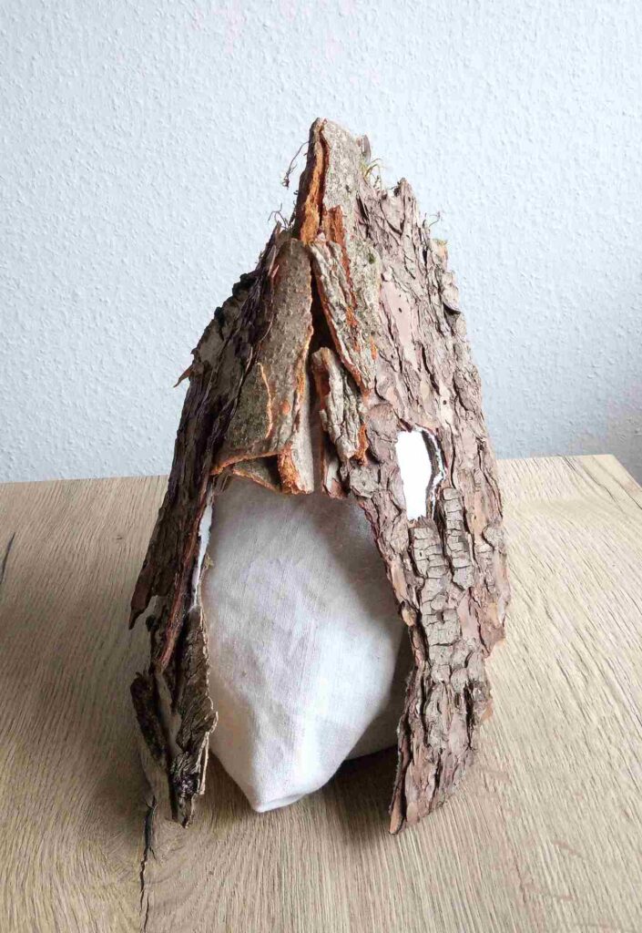 Tobias Funk. 2022. Der Geist der wachsenden Erkenntnis (Ansicht 2). Skulptur, alte Rinde von Birke, Buche, Fichte (Fundort: Marburg), Moos, auf alten Leinenstoff, Restpappe. Verklebung mit Holzleim. Halterung aus verknoteten Bändern alter FFP2 Masken. Innen mit Monogramm gestempelt und Entstehungsjahr versehen. 19,4 x 34,2 x 17,6 cm