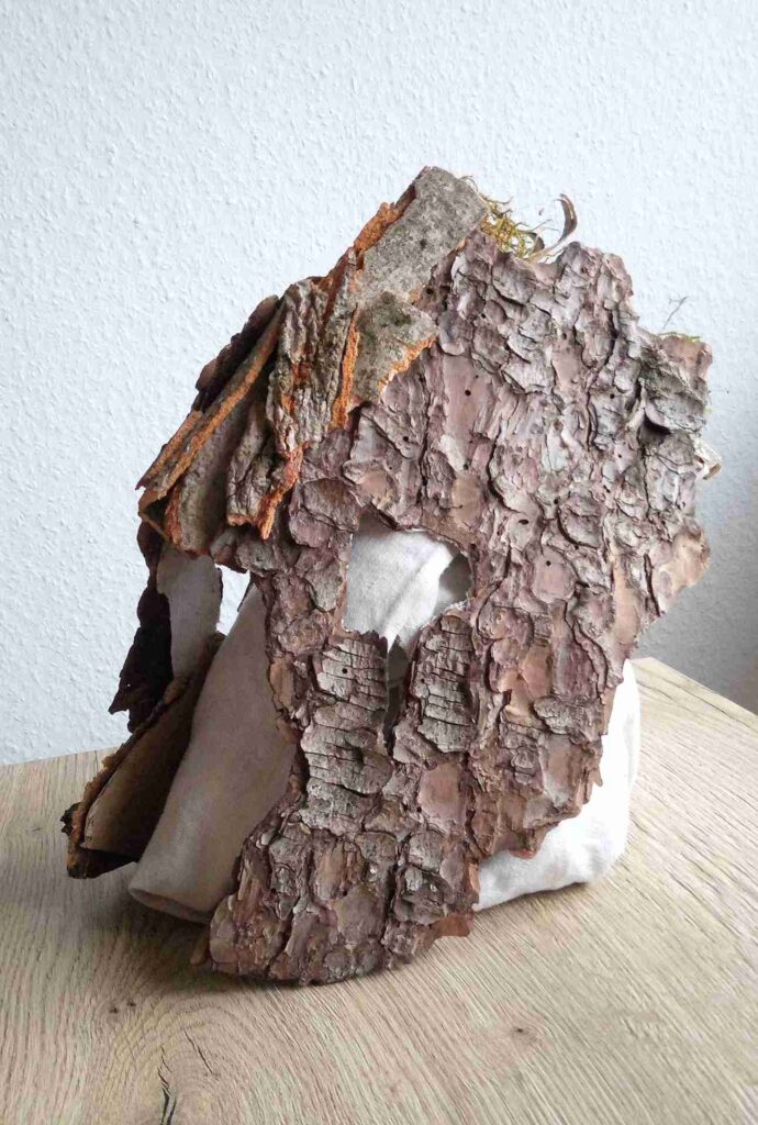 Tobias Funk. 2022. Der Geist der wachsenden Erkenntnis (Ansicht 3). Skulptur, alte Rinde von Birke, Buche, Fichte (Fundort: Marburg), Moos, auf alten Leinenstoff, Restpappe. Verklebung mit Holzleim. Halterung aus verknoteten Bändern alter FFP2 Masken. Innen mit Monogramm gestempelt und Entstehungsjahr versehen. 19,4 x 34,2 x 17,6 cm