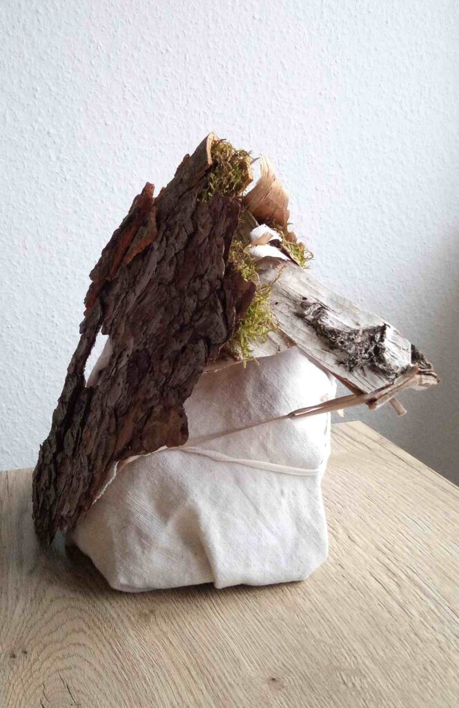 Tobias Funk. 2022. Der Geist der wachsenden Erkenntnis (Ansicht 4). Skulptur, alte Rinde von Birke, Buche, Fichte (Fundort: Marburg), Moos, auf alten Leinenstoff, Restpappe. Verklebung mit Holzleim. Halterung aus verknoteten Bändern alter FFP2 Masken. Innen mit Monogramm gestempelt und Entstehungsjahr versehen. 19,4 x 34,2 x 17,6 cm
