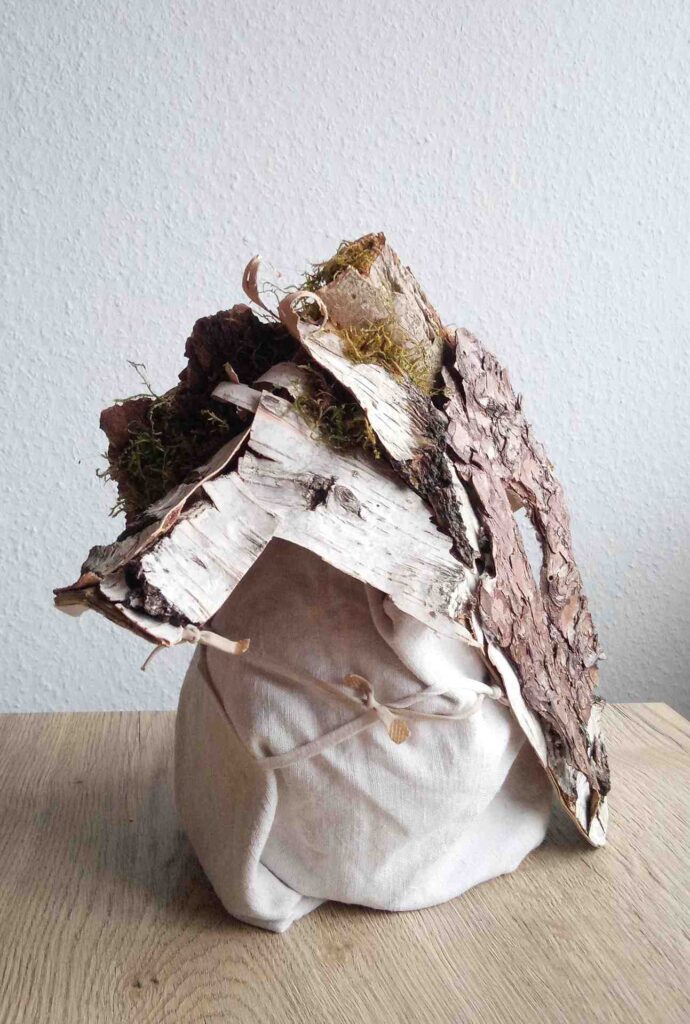 Tobias Funk. 2022. Der Geist der wachsenden Erkenntnis (Ansicht 5). Skulptur, alte Rinde von Birke, Buche, Fichte (Fundort: Marburg), Moos, auf alten Leinenstoff, Restpappe. Verklebung mit Holzleim. Halterung aus verknoteten Bändern alter FFP2 Masken. Innen mit Monogramm gestempelt und Entstehungsjahr versehen. 19,4 x 34,2 x 17,6 cm
