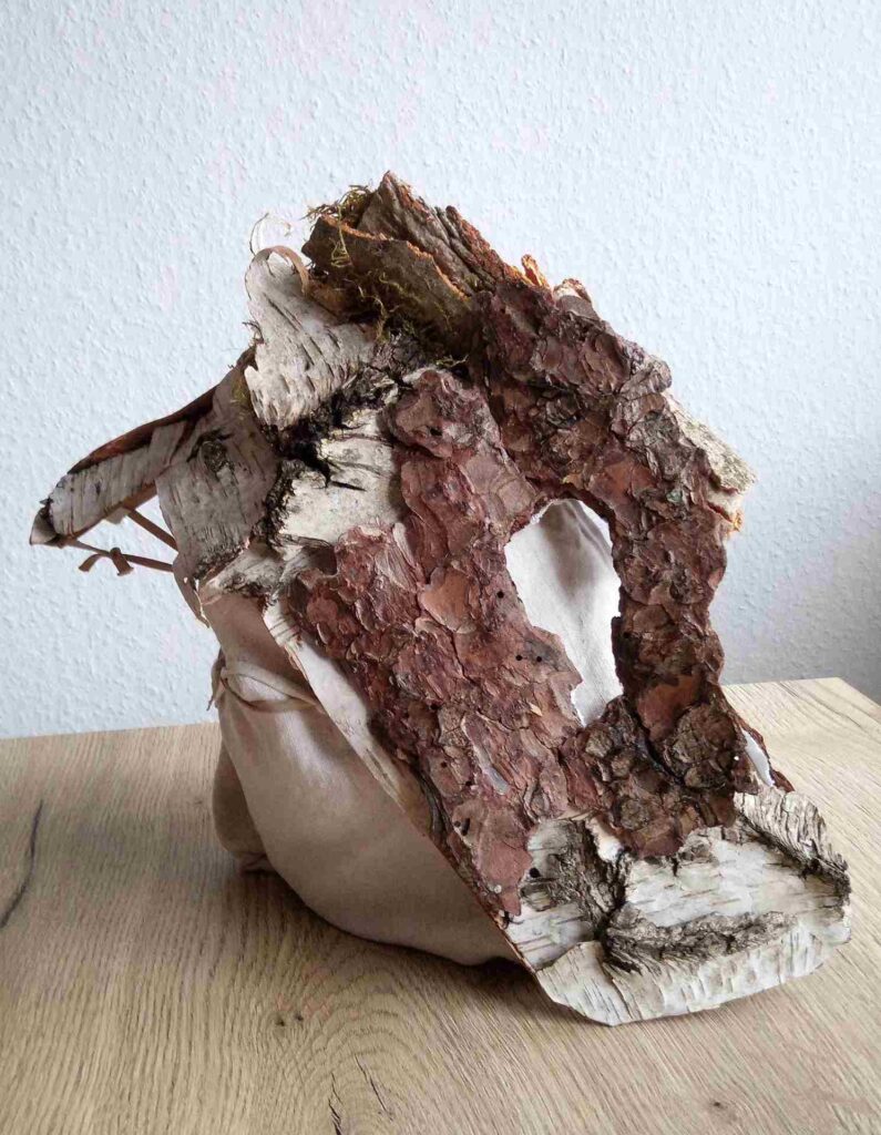 Tobias Funk. 2022. Der Geist der wachsenden Erkenntnis (Ansicht 6). Skulptur, alte Rinde von Birke, Buche, Fichte (Fundort: Marburg), Moos, auf alten Leinenstoff, Restpappe. Verklebung mit Holzleim. Halterung aus verknoteten Bändern alter FFP2 Masken. Innen mit Monogramm gestempelt und Entstehungsjahr versehen. 19,4 x 34,2 x 17,6 cm